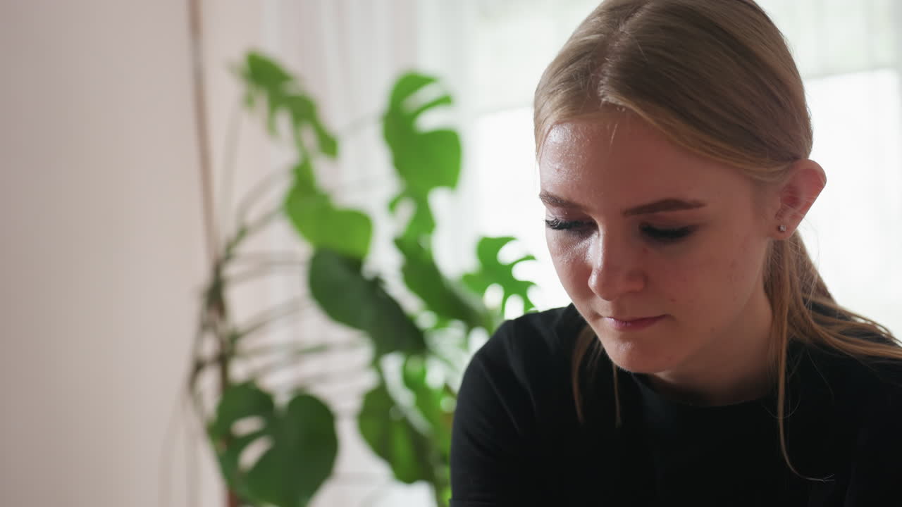 Blonde woman with hair tied back wearing black shirt looking down thoughtfully beside large green plant in softly lit room with white curtains and blurred background