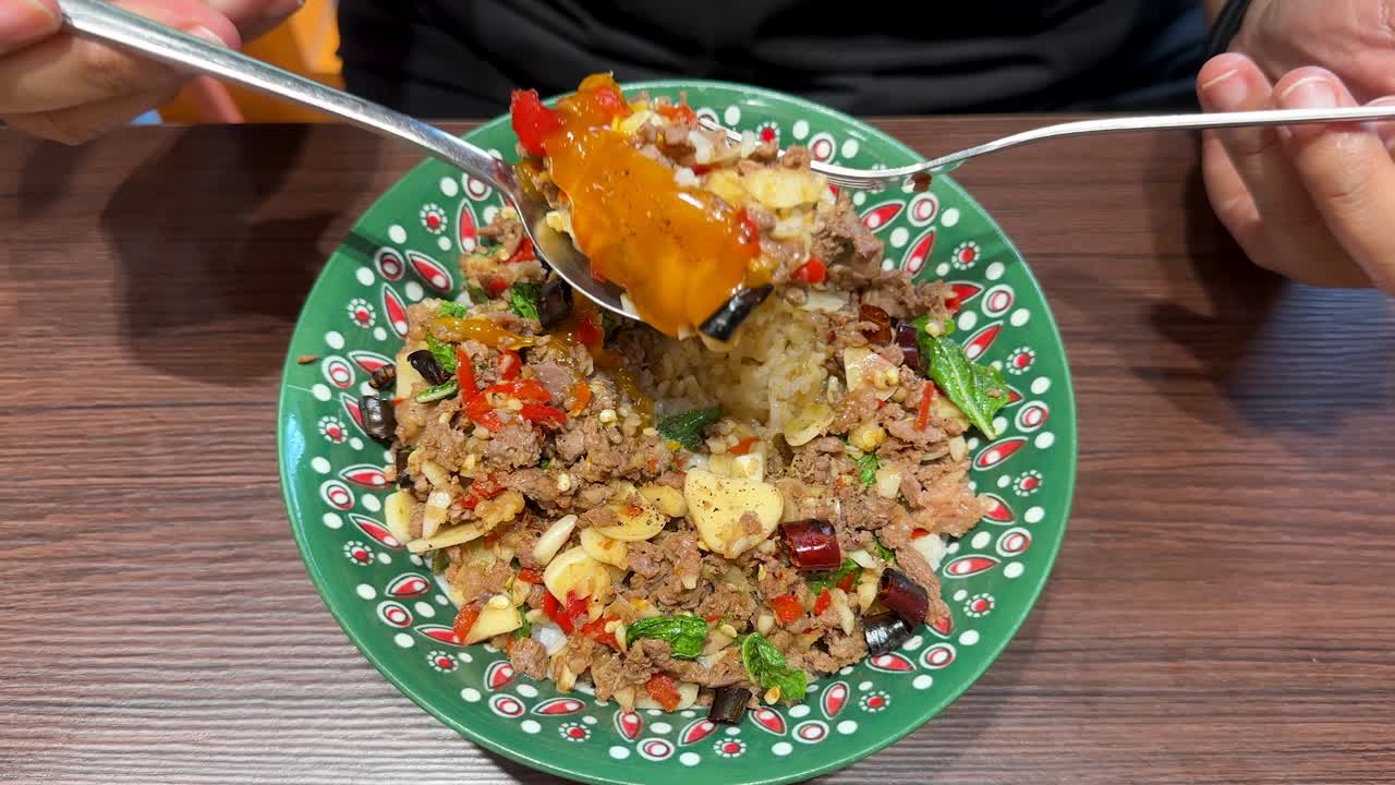 A person uses a spoon and fork to mix a colorful beef, rice, and egg bowl with chili and garlic on a wooden table under bright lighting