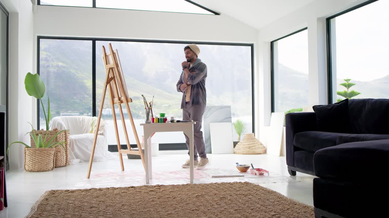 Indian man in beanie painting at easel, mixing paints, drawing inspiration from lake view