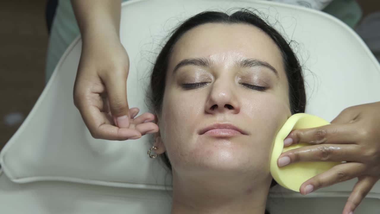 mujer relajada acostada en el salón y teniendo una limpieza profunda tratamiento facial nutritivo. esteticista limpieza rostro del cliente con esponja redonda. atención médica, bienestar y medicina