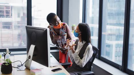 Diverse-female-office-colleagues-wearing-face-masks-discussing-at-modern-office