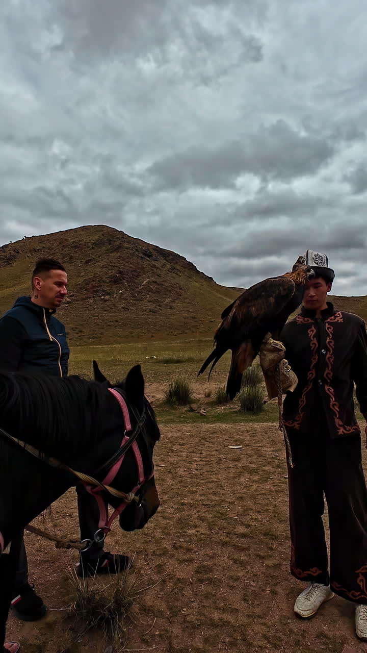 Vertical shot of a man holding an golden eagle on hand on a cloudy day beside a black horse.