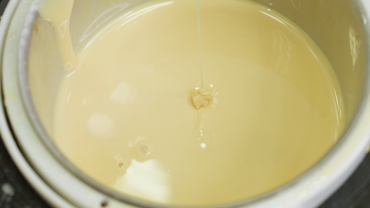 Close up of beautician using wooden stick to stir warm creamy substance inside electric heater, slowly raising stick to observe consistency during preparation in beauty salon environment