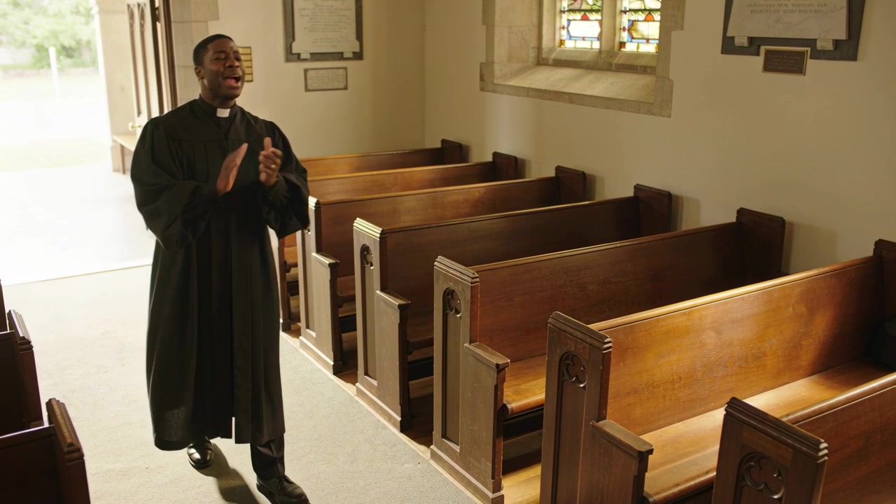 Entering from open door pastor in black robe walking church aisle by pews, clapping leading worship