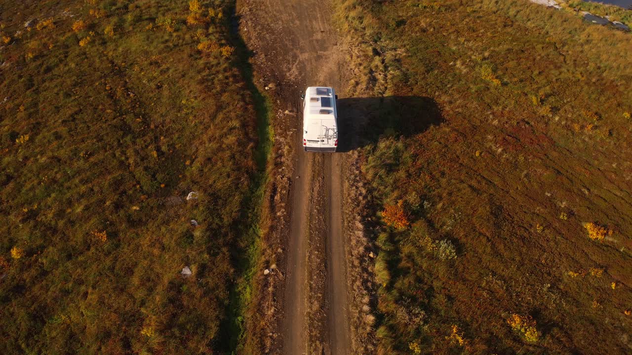 Aerial shot of a white van driving along a dirt road in a rugged autumn landscape