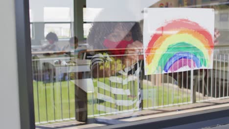 Boy-wearing-face-mask-sticking-rainbow-painting-to-the-window-at-school