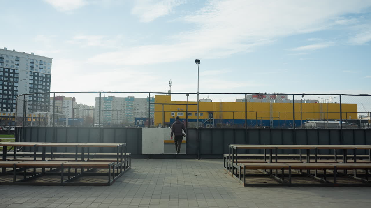 Person walking away from urban sports arena, carrying a soccer ball, amidst a backdrop of vibrant yellow storage containers and city buildings
