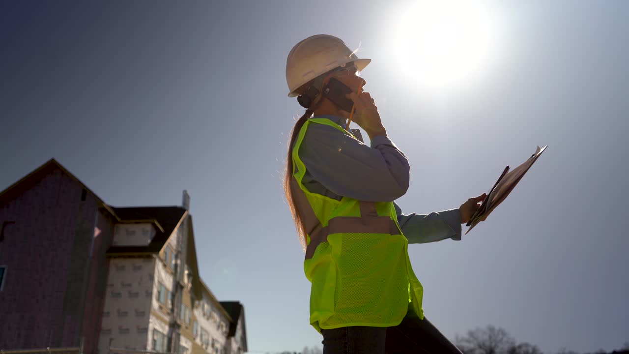 primer plano de gran angular tomado desde el suelo de una ingeniera parada en su lugar hablando en un teléfono inteligente y revisando los planes en el portapapeles con el sol en un cielo sin nubes