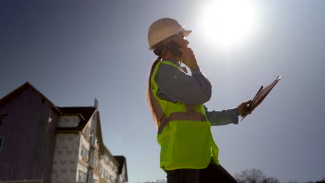 wide angle closeup shot from ground of woman engineer standing in place talking on smartphone and checking with plans on clipboard with sun in a cloudless sky