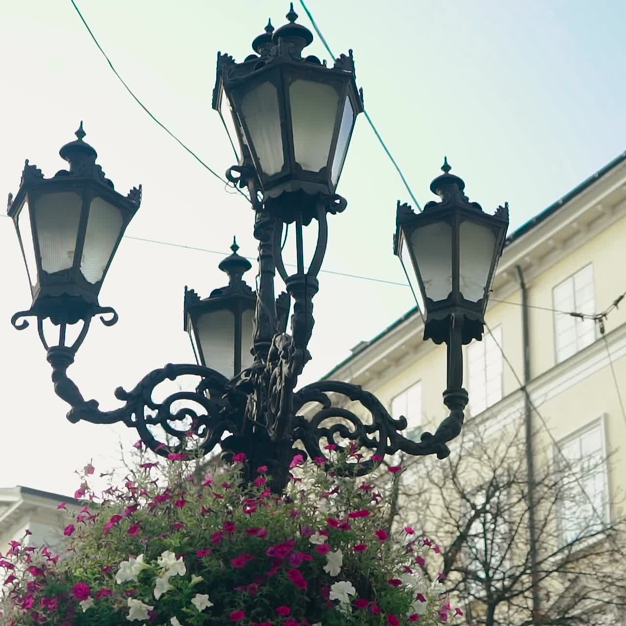 view of the decorative lantern which decorated with flowerpots with purple and white flowers on the background of the building in the city. Camera motion to side