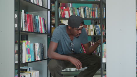 Joven estudiante con camisa informal y gafas de sol en el cuello, en cuclillas en el pasillo de la biblioteca, buscando un libro en la estantería inferior, búsqueda centrada entre lomos coloridos, paredes color turquesa y filas apiladas alrededor