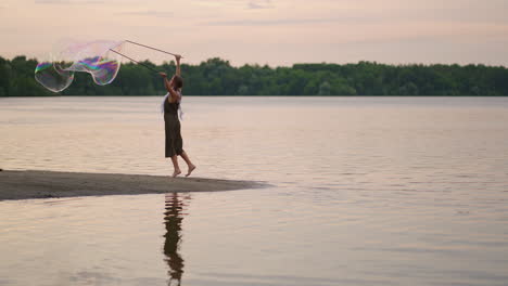 a young female artist shows a soap bubble show blowing up huge soap bubbles on the shore of a lake at sunset. show a beautiful show of soap bubbles in slow motion