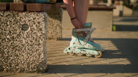 side view of individual seated on bench wearing cyan roller skates while adjusting strap with black bangle on right hand, close by is white sneaker on pavement with sunlight casting shadows