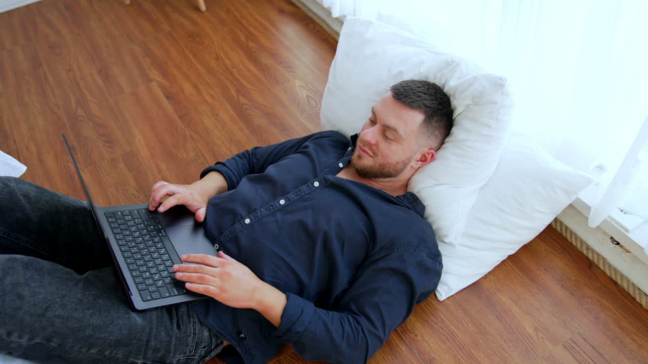 Caucasian male in blue shirt lies on the floor holding feet on the bed. Man works on the laptop set on his belly. Top view.