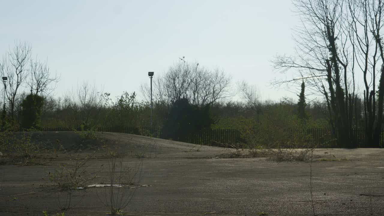 Overview of Broken and Disused Car Park Being Reclaimed by Nature with Weeds and Bushes Growing Out of Tarmac with Old Floodlights and Trees. Post Apocalypctic Setting.