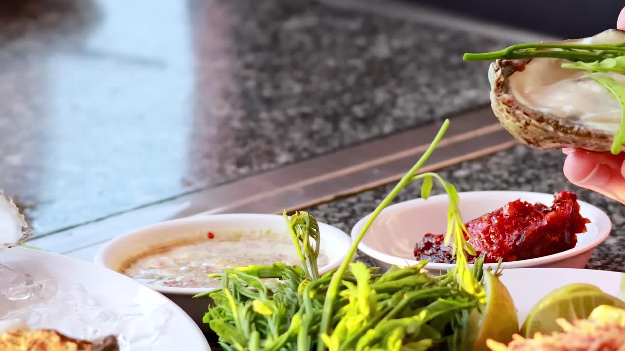 Hands delicately garnish oysters with fresh herbs and a vibrant red sauce on a kitchen counter.