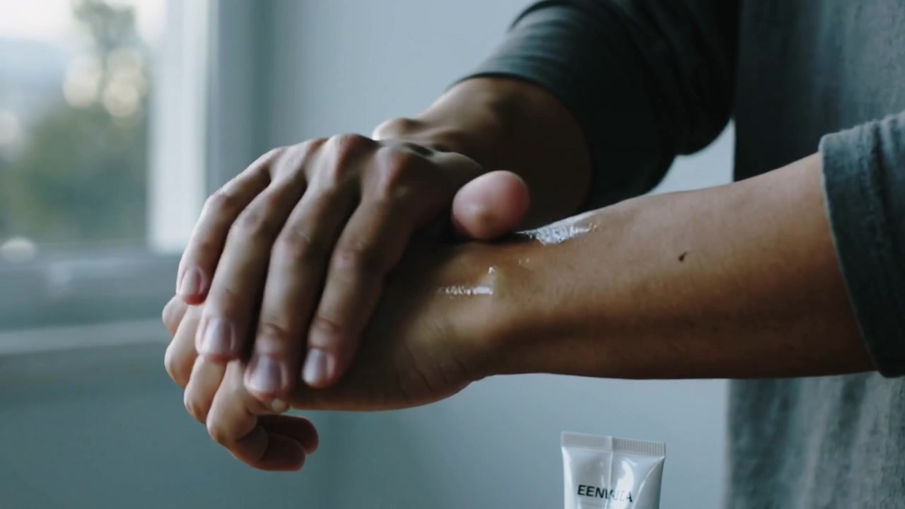 Mand i gråærmet trøje klemmer og påfører creme fra tube på venstre arm ved vinduet, gnider for at fugte.