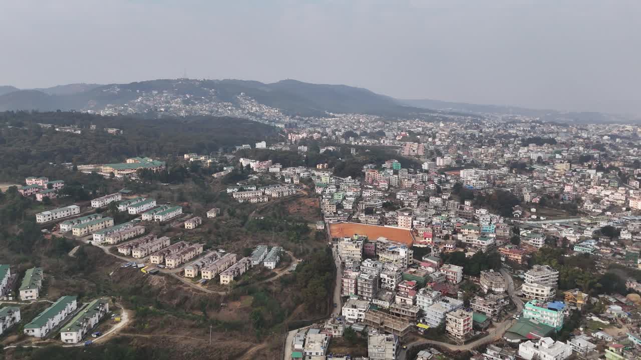 Riprese drone rivelano una vasta città collinare con edifici densamente raggruppati caratterizzati da tetti colorati rossi, verdi e blu immersi tra gli alberi con montagne nebbiose sullo sfondo che mostrano un panorama urbano