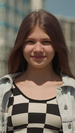 teenage girl with straight hair stands on street of big city on sunny day. teenager in good mood smiles carefree closeup on blurred background