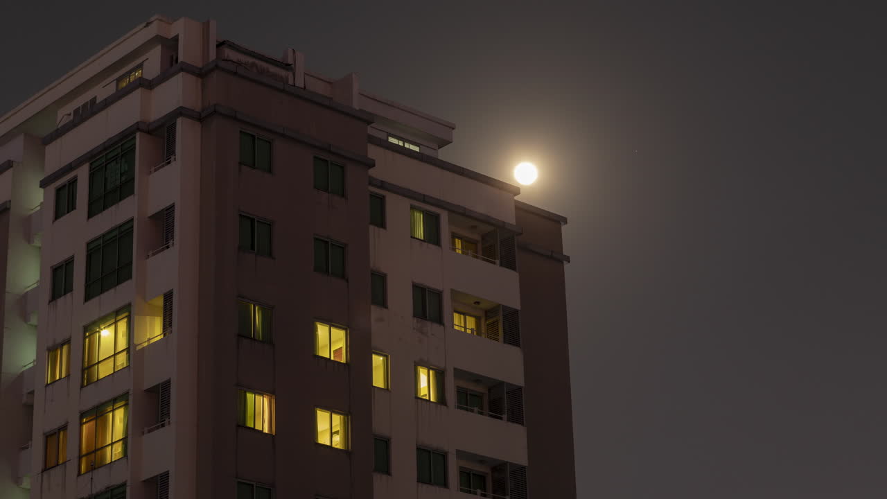 lapso de tiempo de la luna llena que se eleva sobre un edificio residencial mientras las luces se apagan y encienden por la noche