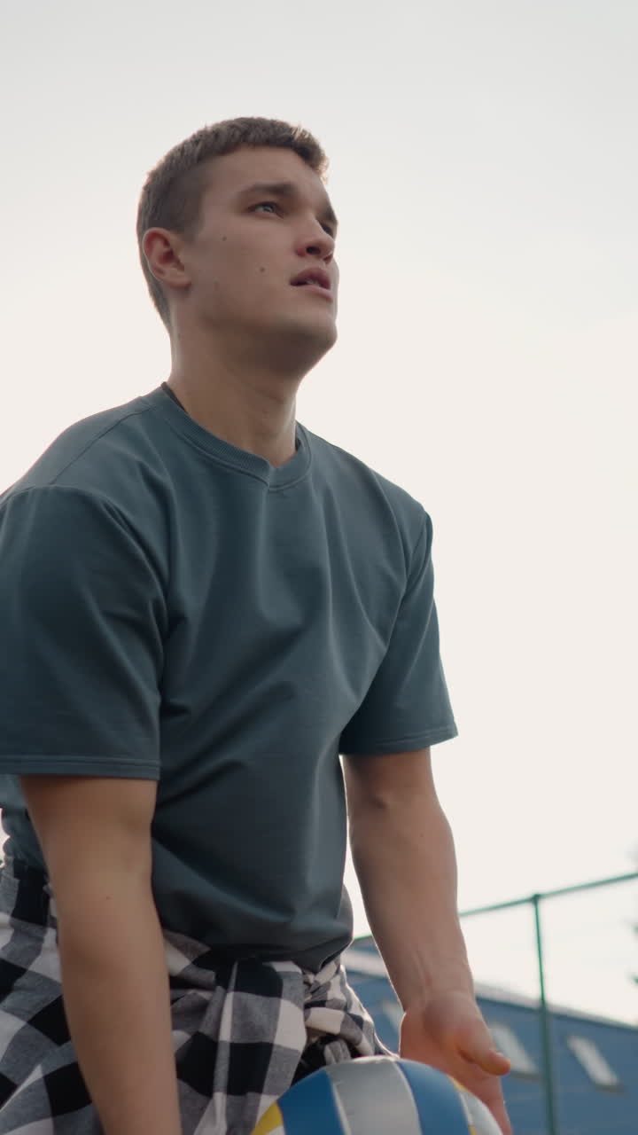 atleta rebotando la pelota de voleibol durante la sesión de entrenamiento, girándola en la mano antes de servir, capturado al aire libre en una arena deportiva con vegetación y edificio en el fondo