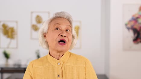 an elderly asian woman in a yellow blouse smiles and makes an ok gesture with her hand in a bright, modern living room. soft, natural lighting and steady camera