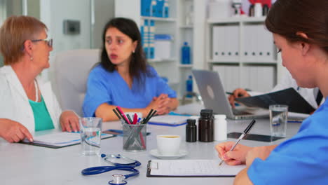 Nurse-writing-on-clipboard-while-profesional-teamworkers-having-medical-meeting