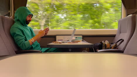 Man-In-Hoodie-Using-Mobile-Phone-While-Traveling-On-Train-With-Laptop-On-Table