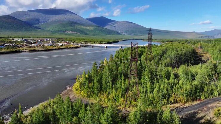Veduta aerea di un fiume che scorre attraverso una valle boscosa con un ponte e tralicci dell'elettricità. La telecamera si sposta a destra per rivelare una strada tortuosa lungo la riva del fiume e dolci montagne verdi