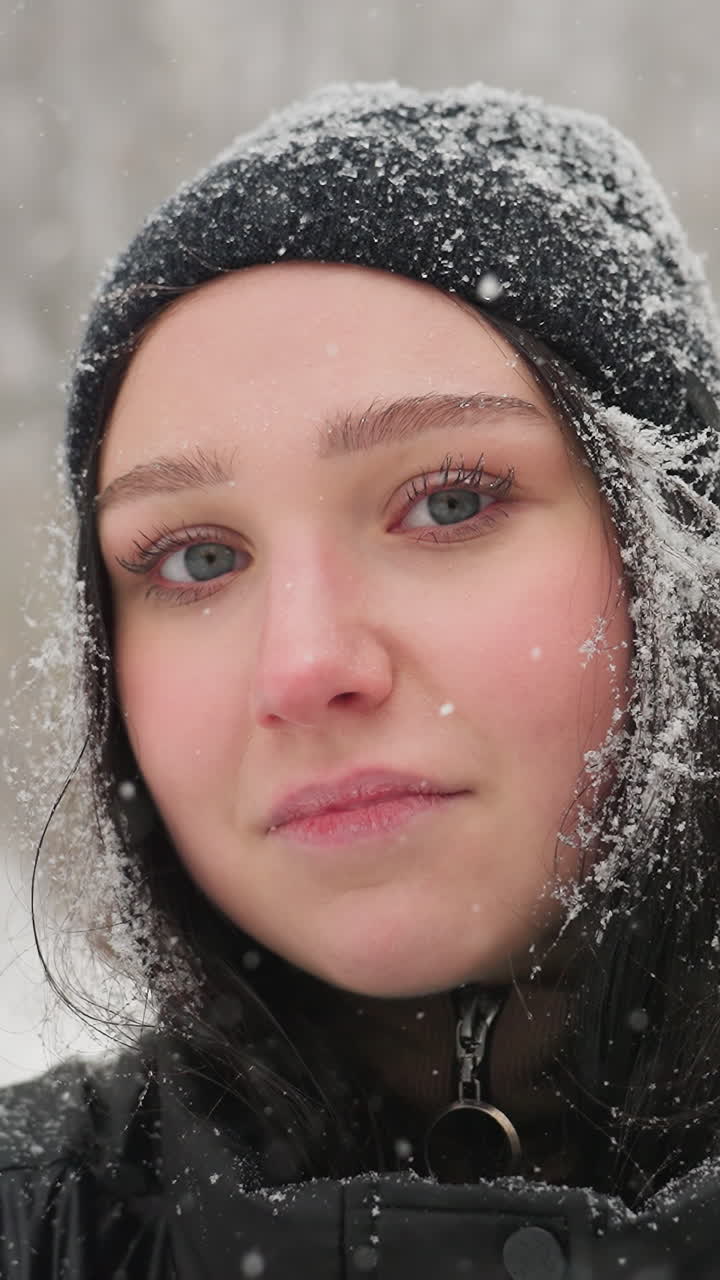 겨울 장면에서 기타를 들고 서있는 어린 소녀, 그녀의 검은 모자와 재에 눈송이가 쉬고, 평온하고 반사적인 표정으로 카메라를 직접 쳐다보고 있습니다.