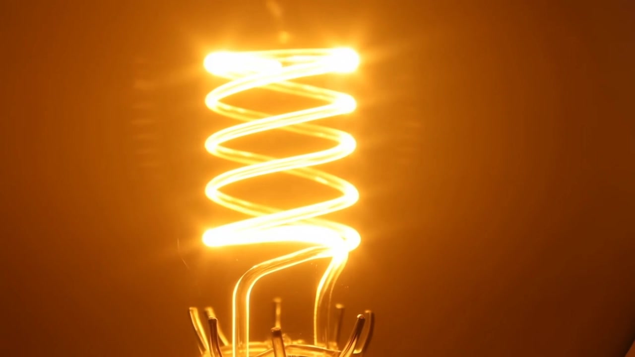Lampada a filamento a spirale che si illumina, emettendo una luce ambra calda in una lampadina di vetro a casa mentre la potenza aumenta, sbocciando.