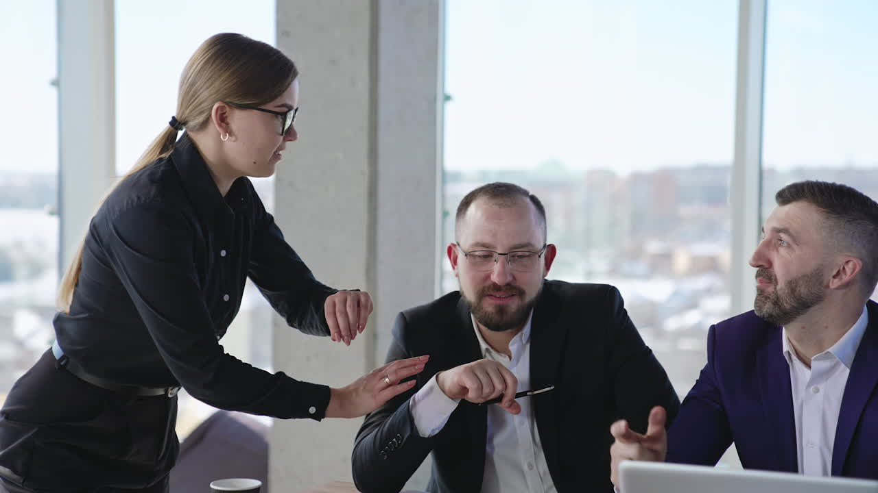 Two man sitting at the desk listen to a woman standing beside. Lady in black clothes looks at her watch and tells something to the listeners. Cityscape in blur at backdrop.