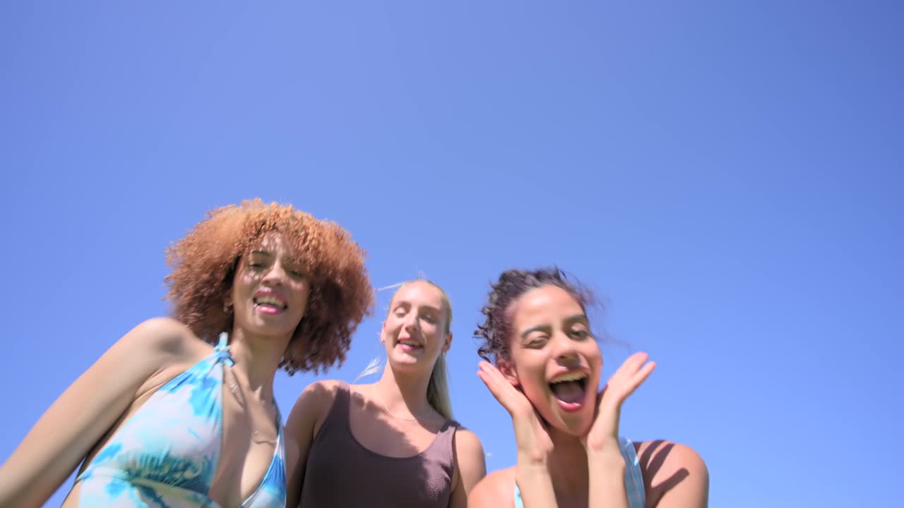 Amiche multietniche che si avvicinano a una telecamera bassa, posano in costume da bagno in giardino, con il sole che provoca un bagliore sull'obiettivo.