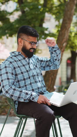 Indian-happy-man-working-on-laptop-celebrate-success-win-money-sitting-on-urban-street-in-city