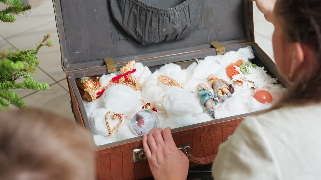 Back view of woman opening vintage box filled with Christmas ornaments surrounded by soft white cotton, festive cozy indoor atmosphere with decorated Christmas tree glowing in background