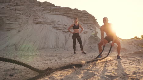 athletic man on exercise around the sand hills at sunset hits the rope on the ground and raised the dust.