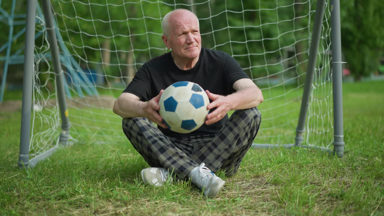 An elderly man sits cross-legged near a goal post, gently toggling a soccer ball in his hands while looking around, with a blue pole in the background and trees