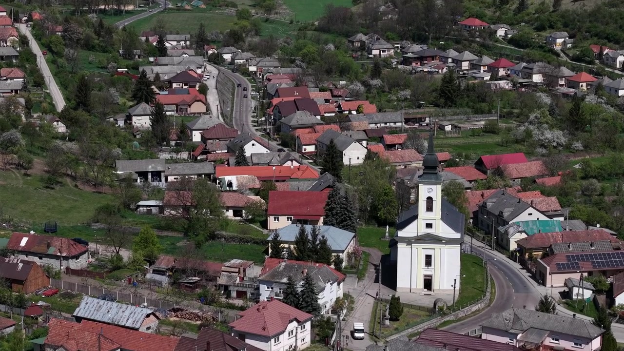 Tradiční evropská vesnice se rozprostírá po zelených kopcích, výrazný bílý kostel a obytné domy ukazují klidnou venkovskou komunitu.