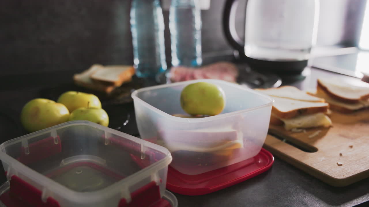 Caregiver places sandwich into lunch container while organizing kitchen items during morning meal prep, with apples, plastic boxes, and cutting board visible, showing daily food preparation routine at home