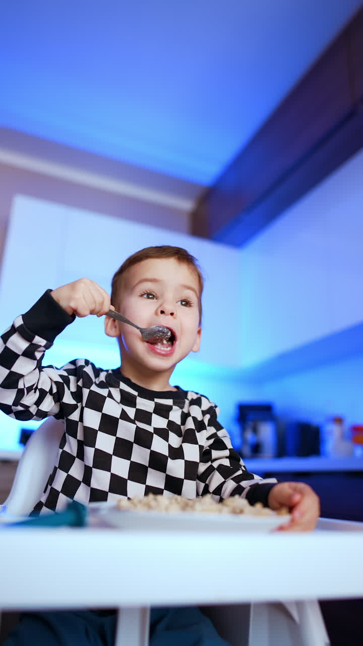 Cute Caucasian toddler sitting at high chair eats porridge. Baby takes a spoon to his mouth and tilts his head. Low angle view. Vertical video.