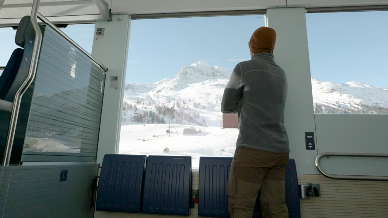 Young caucasian man standing next to a train window and looking outside to snow covered mountains during a sunny winter day