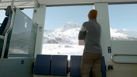 Young-caucasian-man-standing-next-to-a-train-window-and-looking-outside-to-snow-covered-mountains-during-a-sunny-winter-day