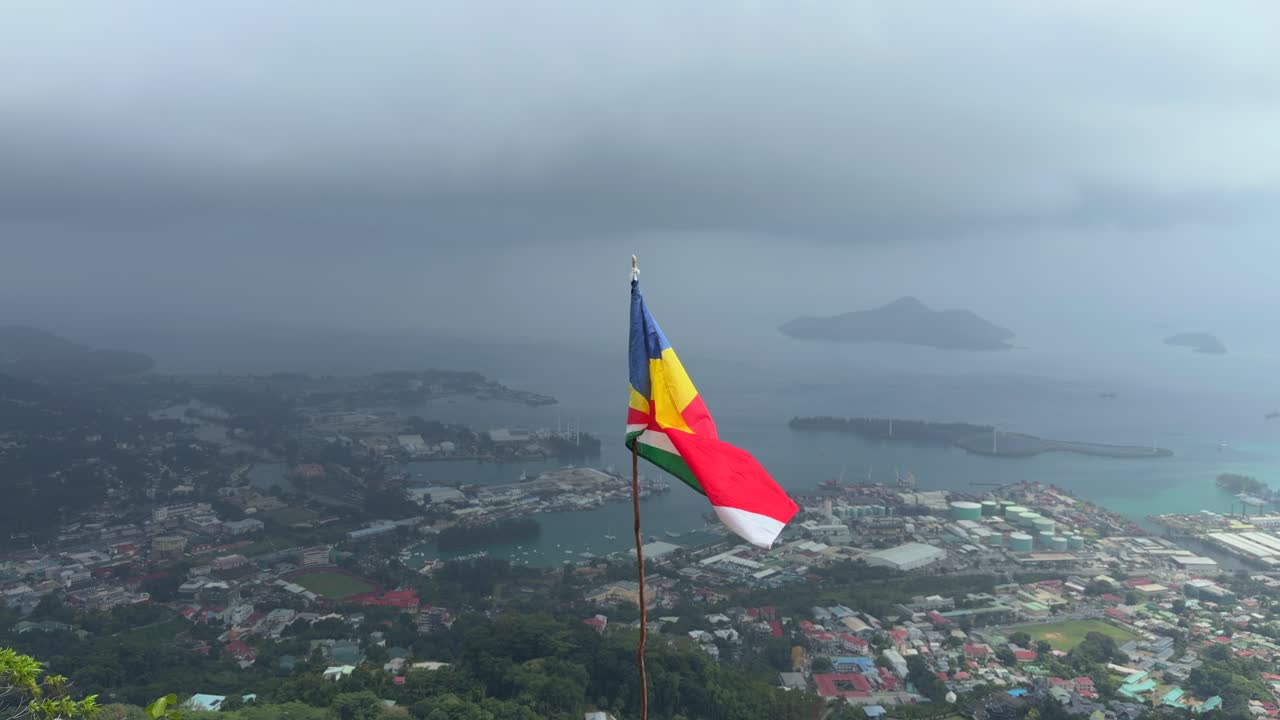 Bandiera delle Seychelles che sventola sopra la collina, sotto la città di Victoria e l'area portuale, Mahe, Seychelles 25 fps 002