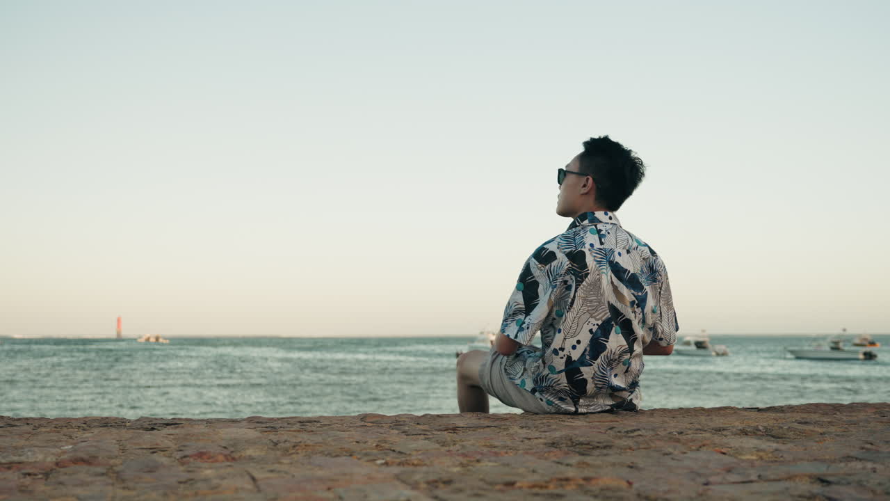 Asiatisk mand sidder på en sten ved stranden og slapper af på ferie med solbriller.