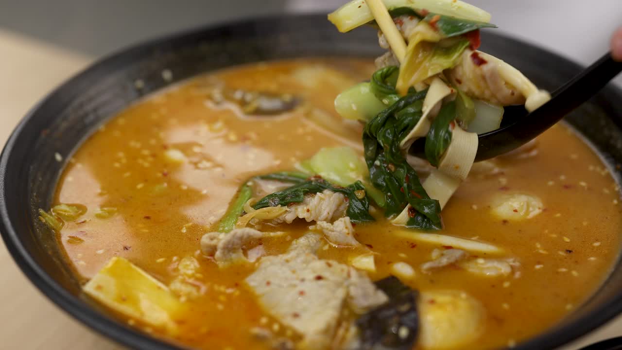 Chopsticks and spoon lift pork, vegetables, and noodles from steaming spicy hotpot under soft lighting