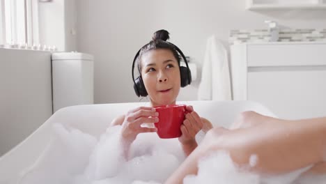 video de una mujer biracial sonriente con auriculares y una taza roja de té en la bañera en un baño de burbujas