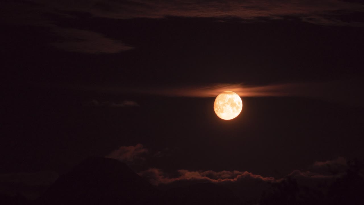 Time lapse of a golden moon moving from left to right side of the frame, surrounded by clouds and a small mountain in the bottom of the frame.
