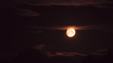 time lapse of a golden moon moving from left to right side of the frame, surrounded by clouds and a small mountain in the bottom of the frame.