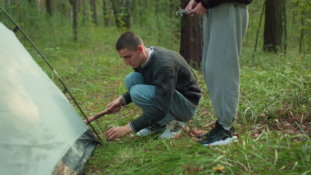 man squats beside tent receiving peg from woman while holding wood stick preparing to hammer peg into forest ground to secure tent during outdoor camping setup surrounded by trees
