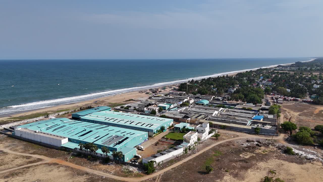 Drone footage captures a modern factory building situated on the shoreline. The industrial development is a stark contrast to the nearby coastal village and serene beach
